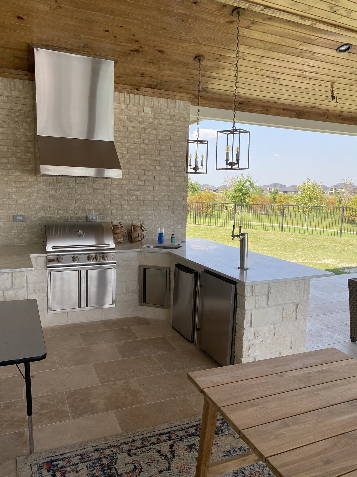 Outdoor kitchen with stainless vent hood and pendant lights under a tongue-and-groove ceiling